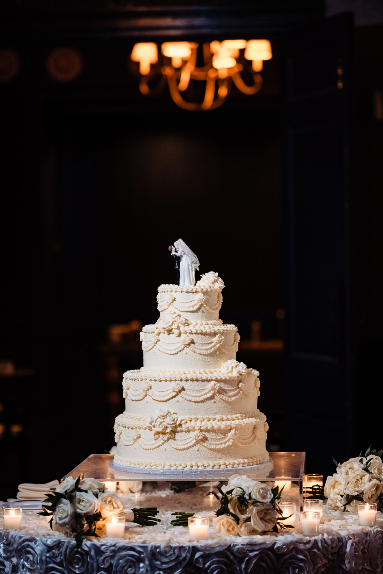 Wedding cake illuminated by soft ambient lighting, with delicate details and a figurine cake topper standing out against a darkened background.