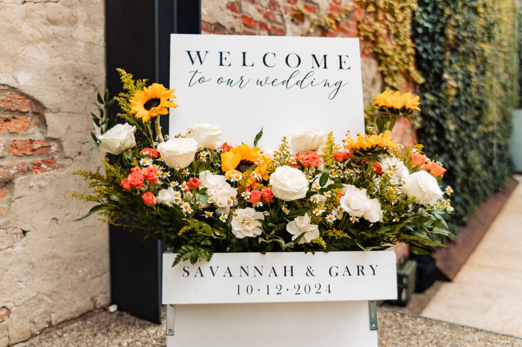 Welcome sign at the wedding entrance with a floral arrangement of sunflowers and white roses, displaying the names Savannah & Gary and their wedding date.