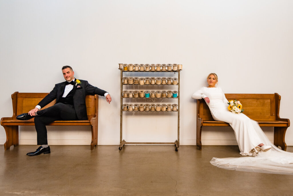 Groom sitting on a bench in a tuxedo, striking a relaxed pose with one leg crossed, while the bride sits gracefully on a nearby bench.