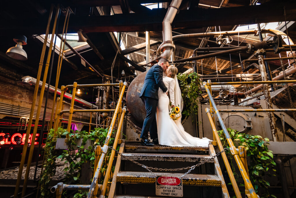 Bride and groom sharing a kiss on a staircase surrounded by industrial decor, with a 'Danger' sign nearby in their Global Dye Works Wedding reception