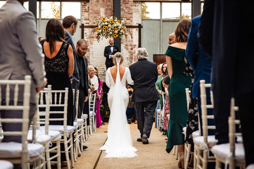 Bride walking down the aisle with her father, captured from behind, as guests stand to witness the ceremony