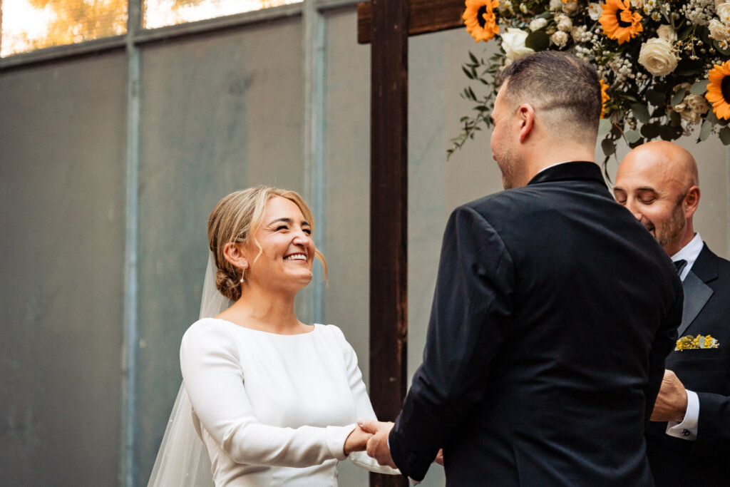 Bride and groom exchanging vows at the altar, with a floral backdrop and their hands clasped