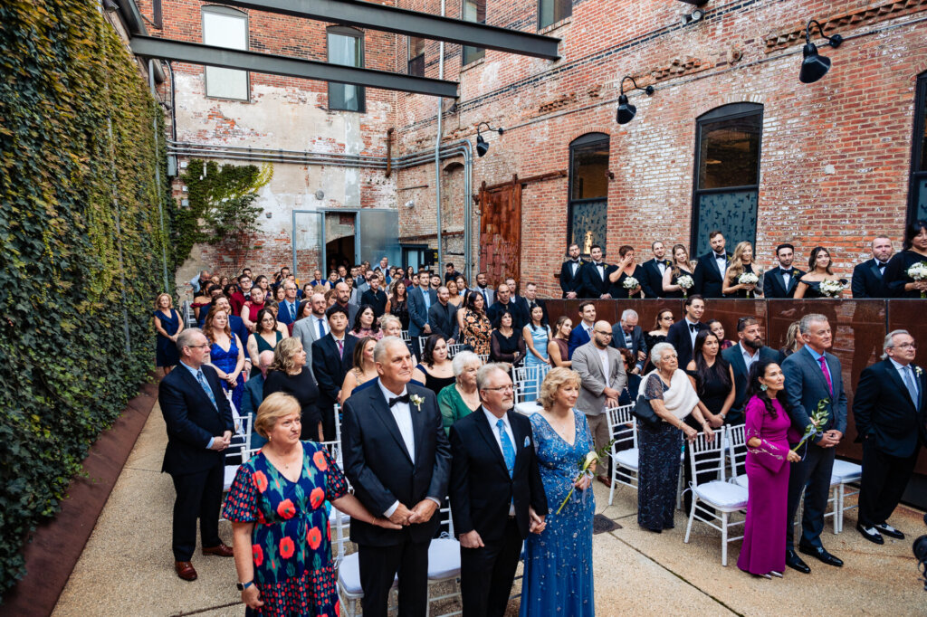 Wide shot of the guests at the wedding ceremony, standing and watching the bride and groom
