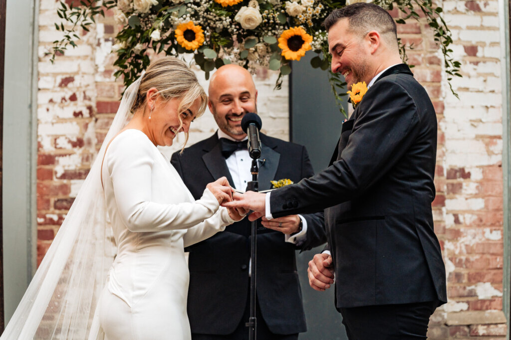 Close up of the bride and groom exchanging rings as they stand at the altar, with the officiant in the background.