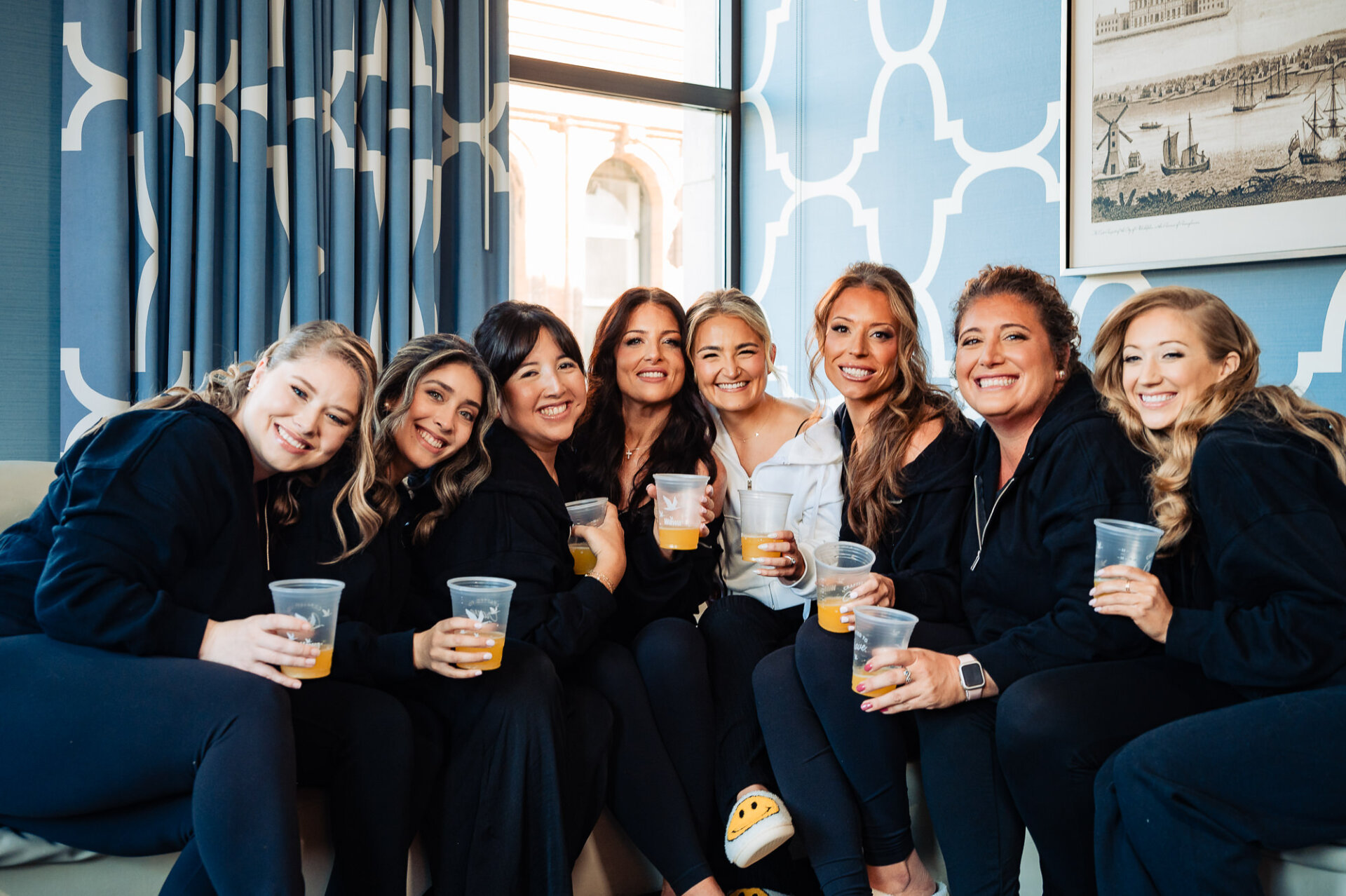 Bride and bridesmaids sitting on a sofa, smiling in matching robes, and holding glasses of orange juice.