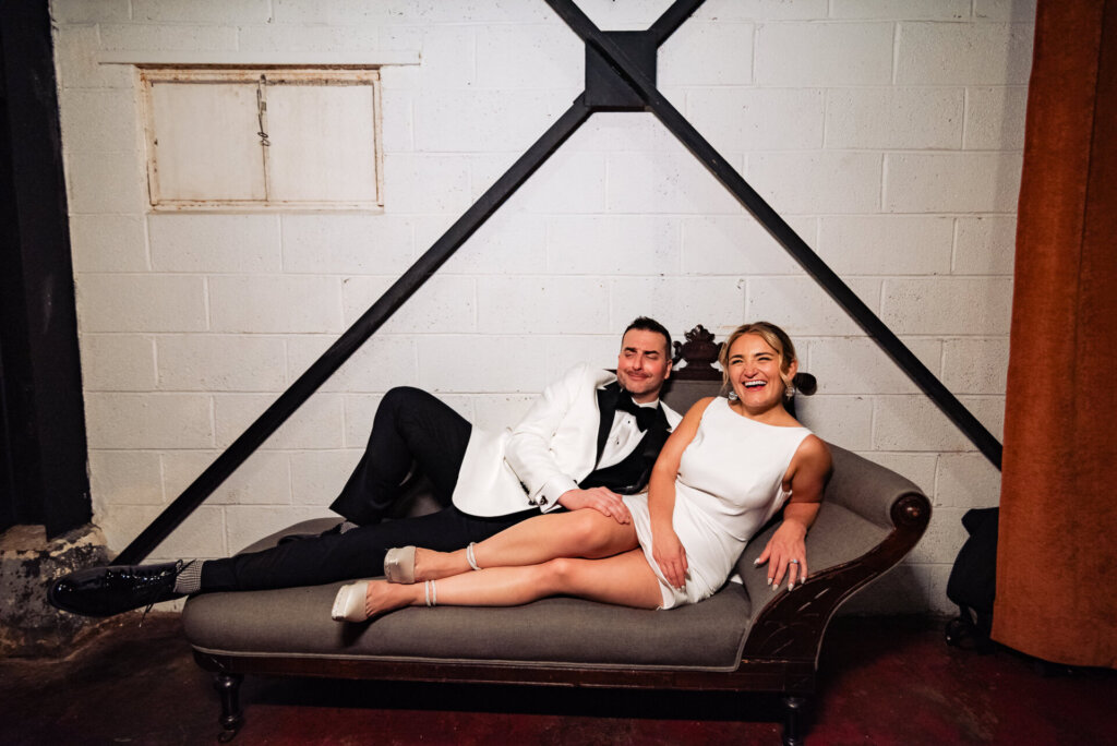 Bride and groom lounging together on a chaise lounge, smiling and enjoying a relaxed moment