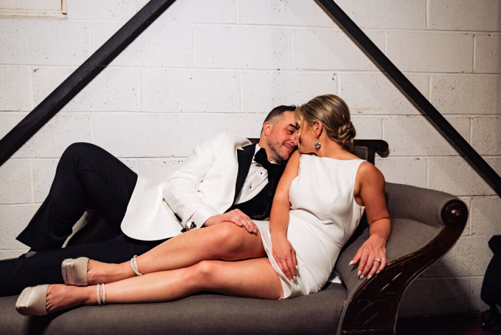 Bride and groom playfully posing on a couch, with the bride leaning on the groom.