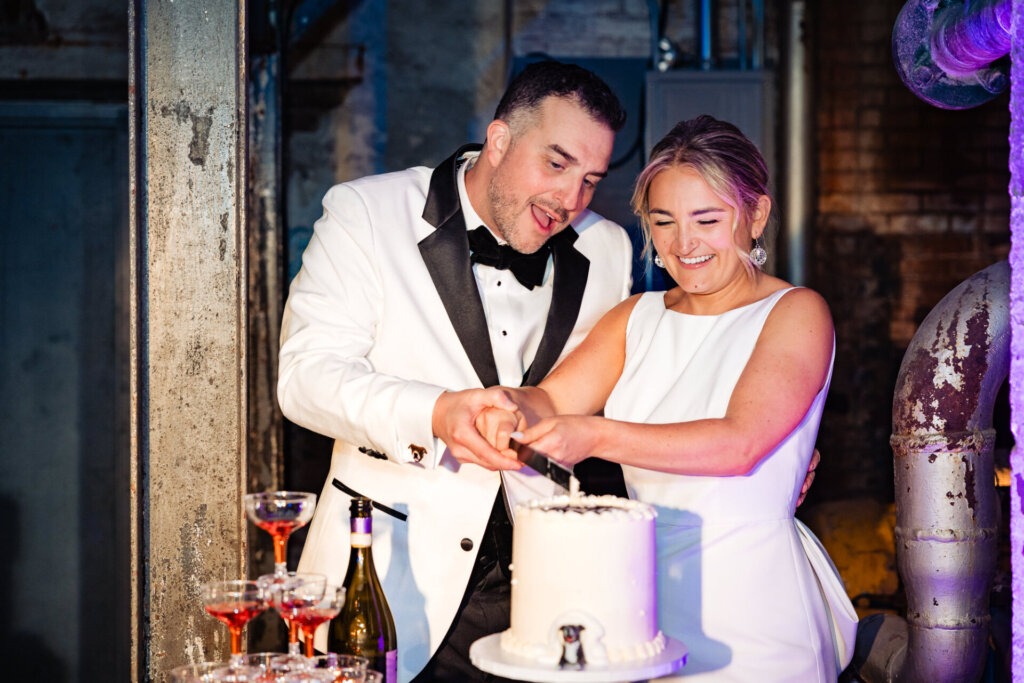 Bride and groom cutting the wedding cake as guests look on, celebrating the moment.