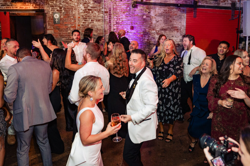 Wedding guests dancing and mingling on the reception floor, with the bride and groom in the crowd.