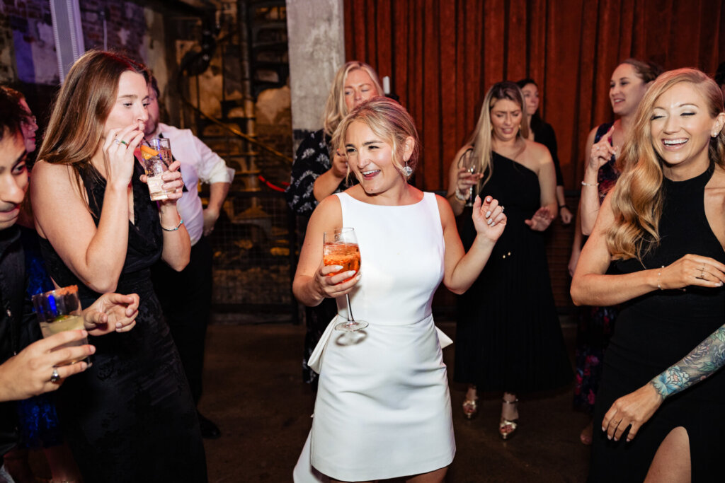 Bride holding a drink, smiling and dancing joyfully alongside guests at the wedding reception.