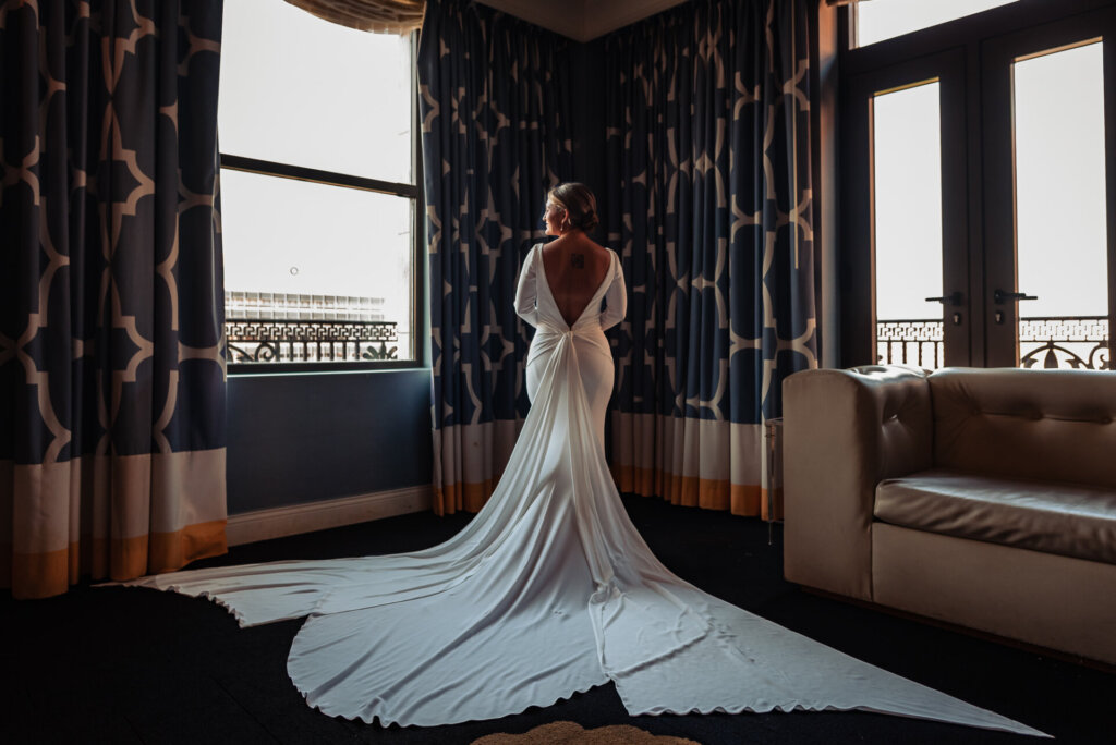 Bride in a long-sleeved wedding gown with a dramatic train, standing in a room with patterned curtains and looking out the window