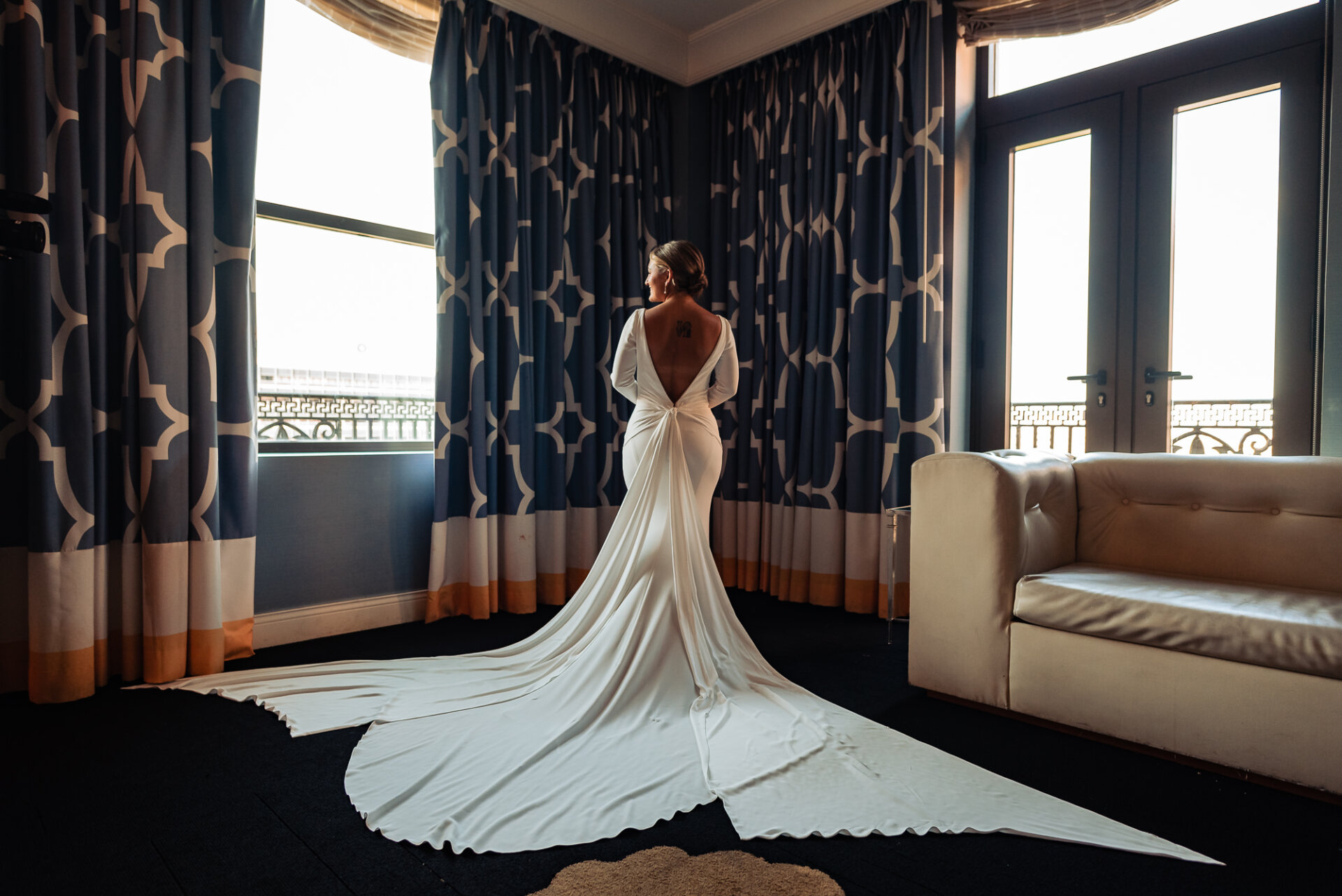 Bride in a fitted white gown with a long train, standing by a window in a softly lit room, looking over her shoulder