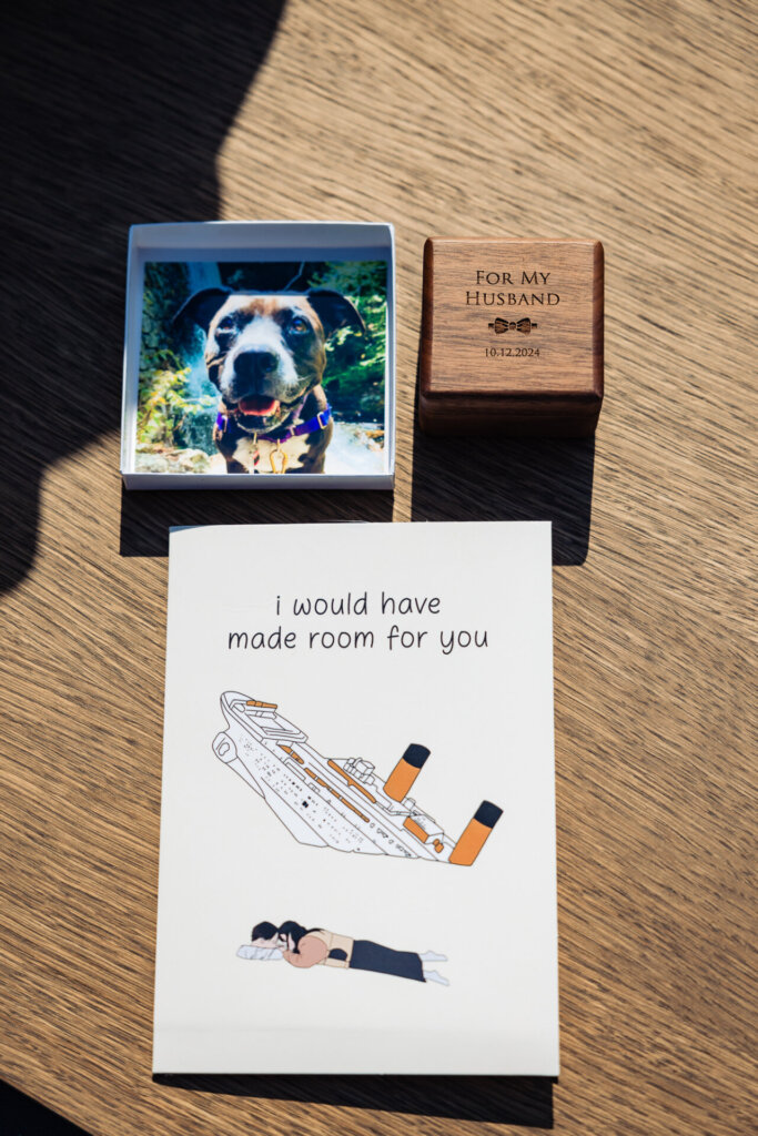 Flat lay featuring a humorous wedding card with Titanic illustration and text, a photo of a dog in a white frame, and a wooden box engraved with 'For My Husband' and the date 10-12-2024.
