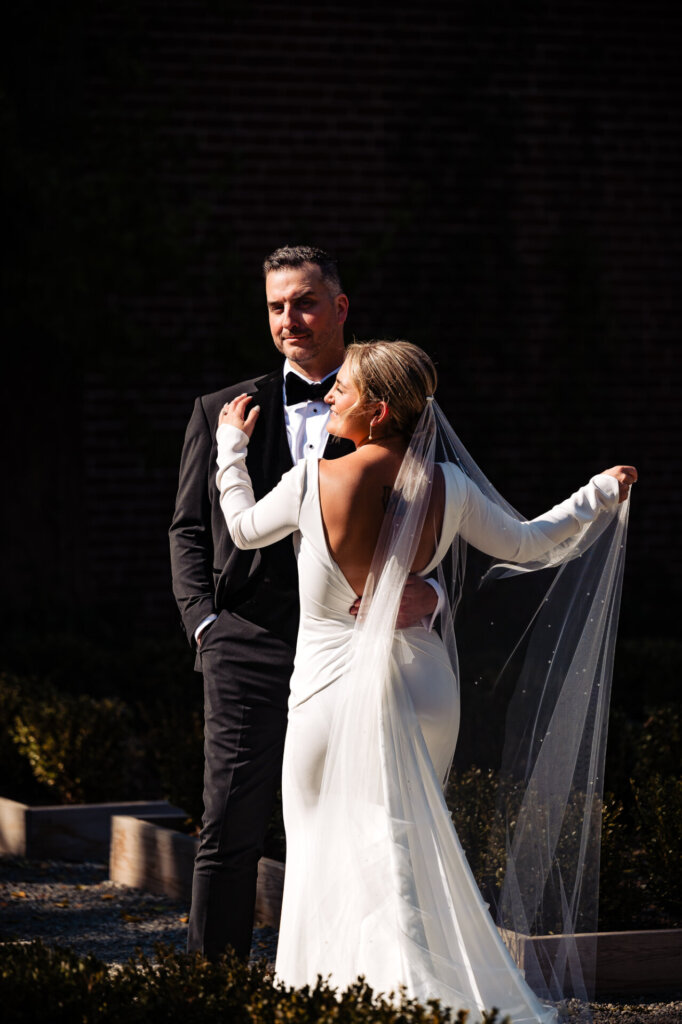 Bride and groom posing together, with the groom holding the bride's waist as she holds her veil, facing away from the camera.