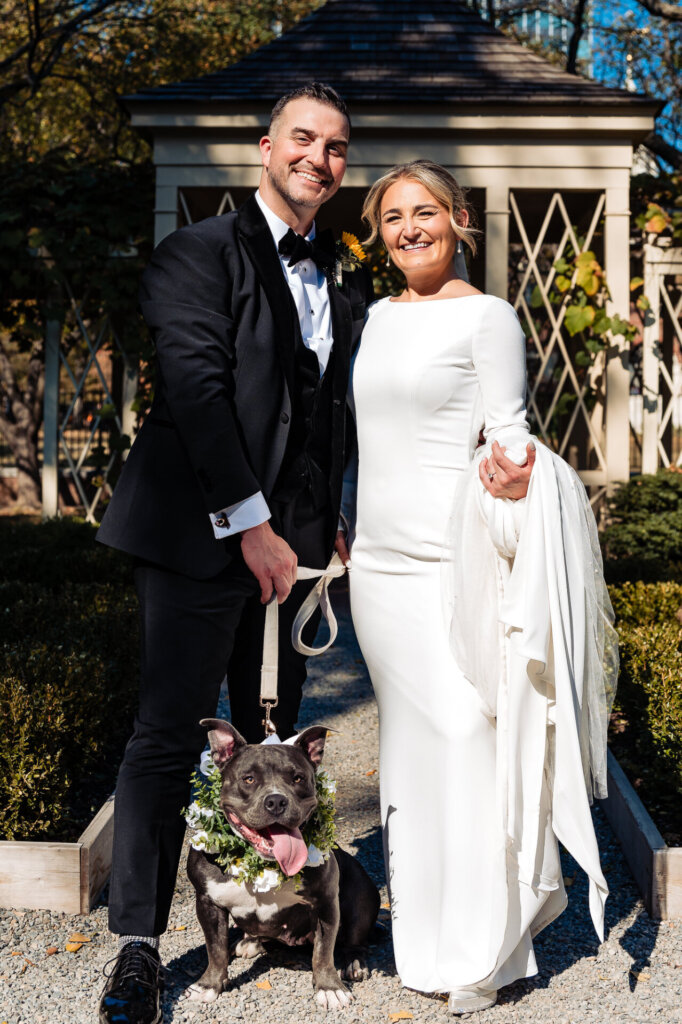 Bride and groom posing together while holding their dog's leash, smiling in a joyful moment.