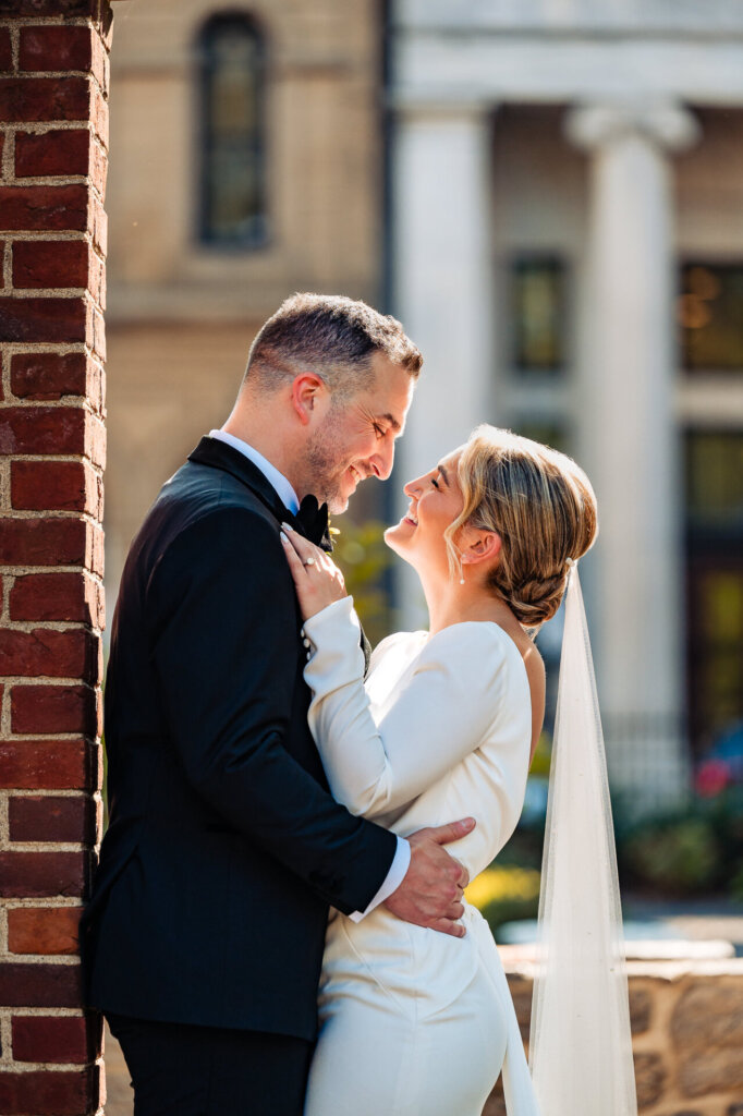 Groom leaning on a brick wall as the bride leans into him, facing each other and smiling, with the groom's hand resting on her waist.