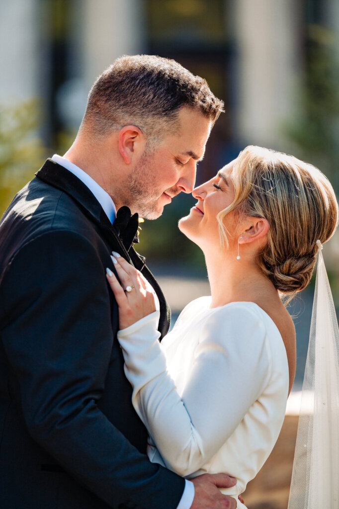 Groom leaning on a brick wall as the bride leans into him, both smiling and facing each other, with the groom's hand gently resting on her waist.