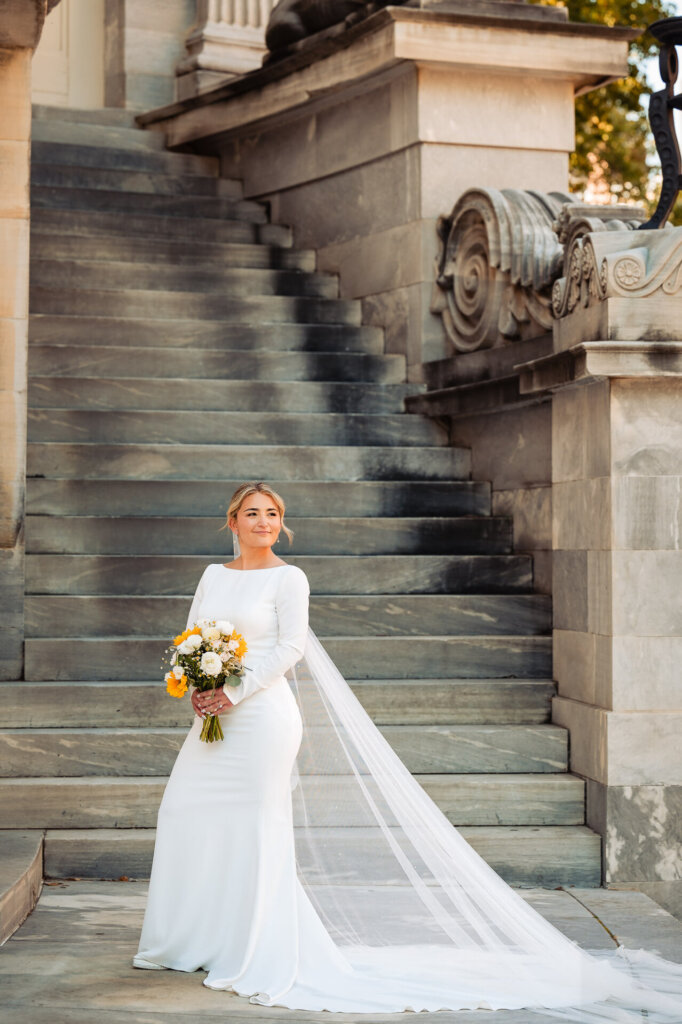 Bride standing on stairs, wearing her wedding gown and holding her bouquet of flowers.
