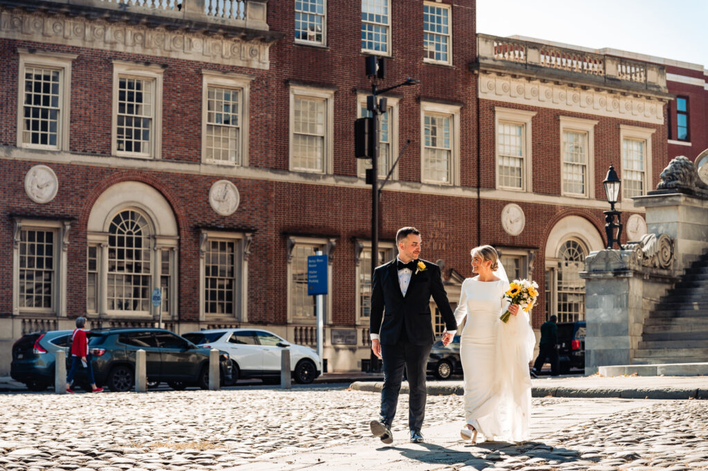 Bride and groom walking hand in hand on a cobblestone street, with a historic building in the background