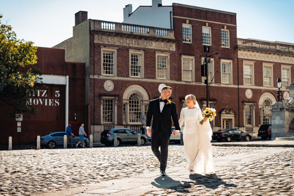 Bride and groom strolling together, smiling, with a bright sunny backdrop of city buildings.