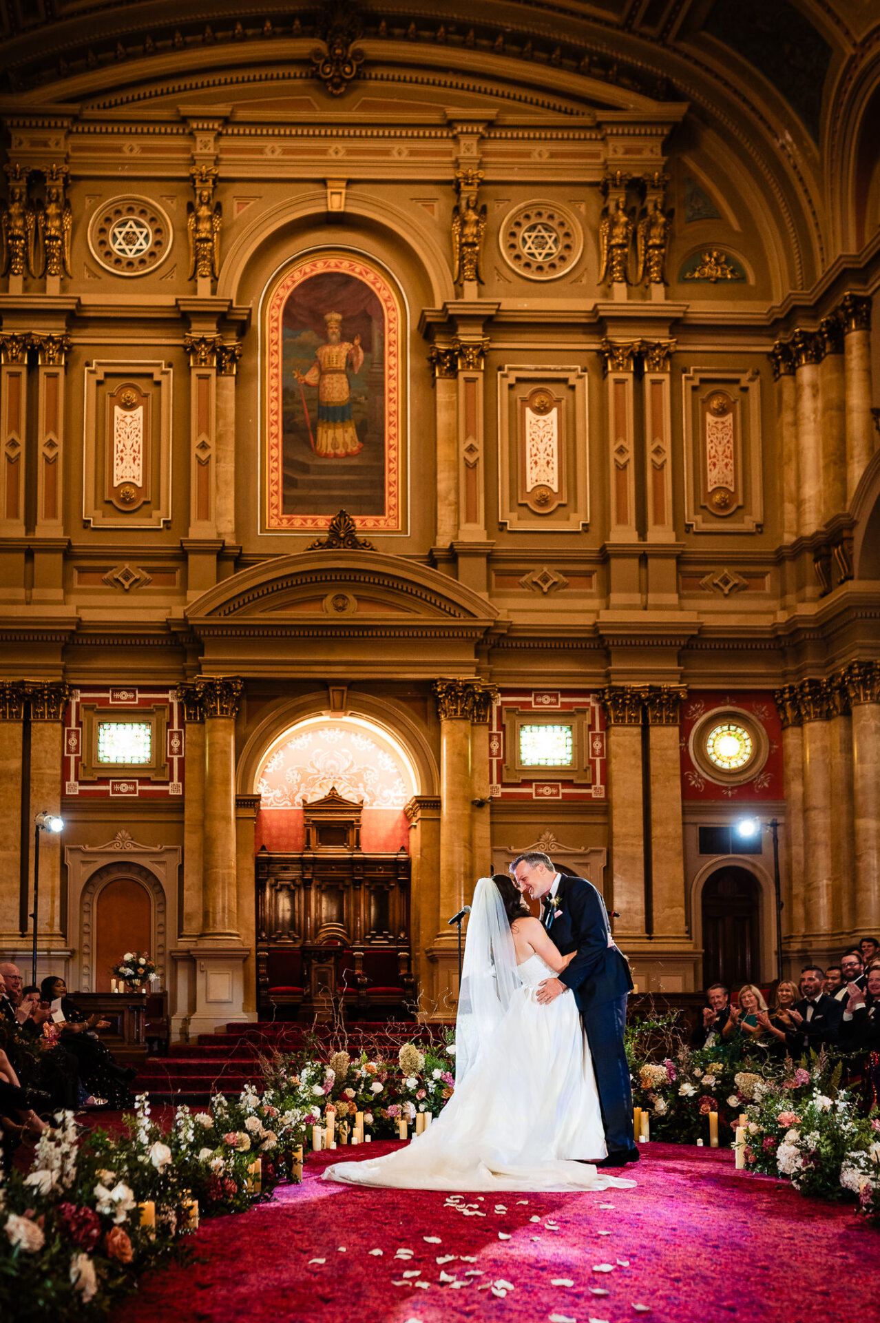 Bride and groom sharing their first kiss at the altar, surrounded by floral arrangements and candlelight in an ornate ceremony hall.