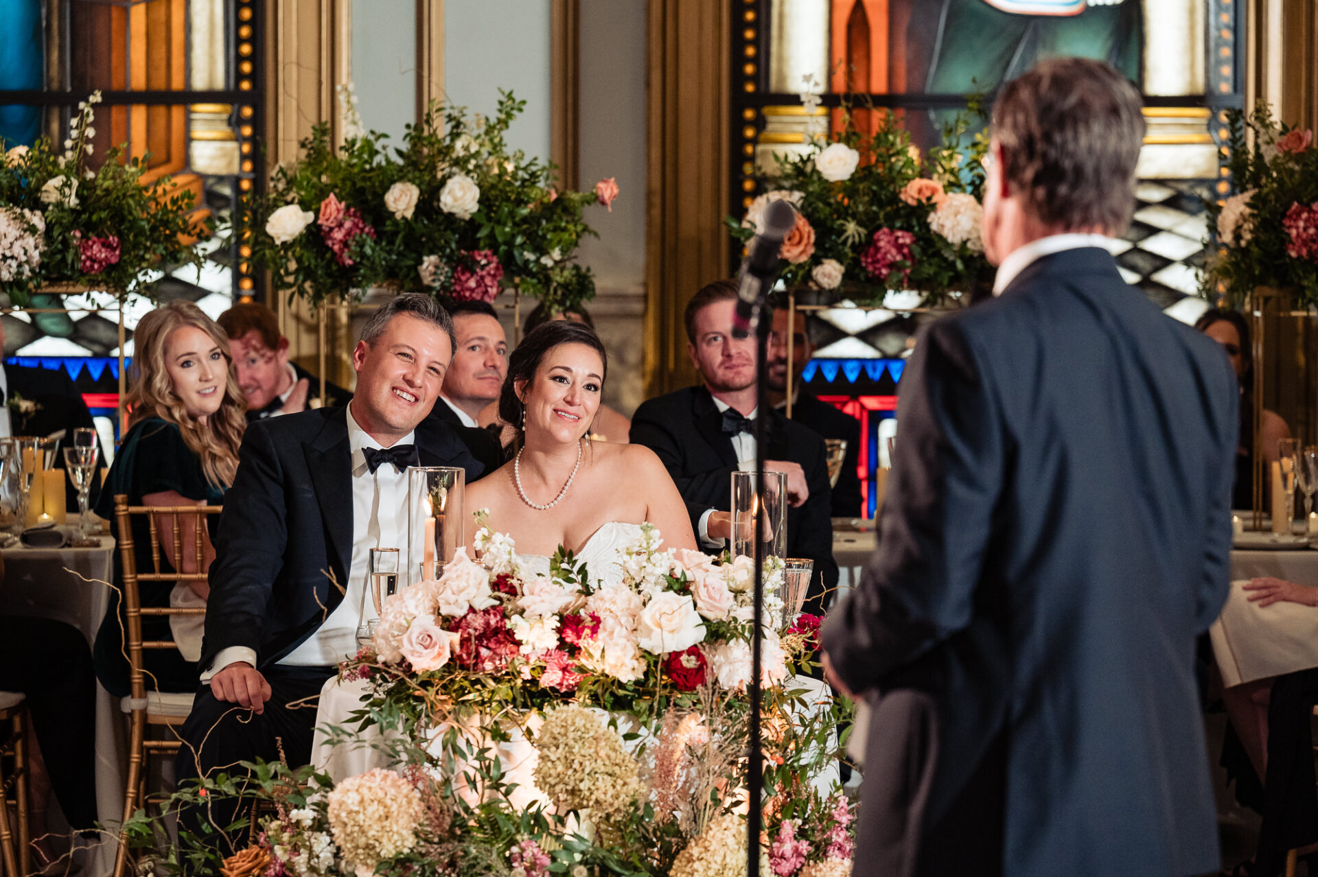 Bride and groom smiling at the head table during a reception speech surrounded by floral arrangements