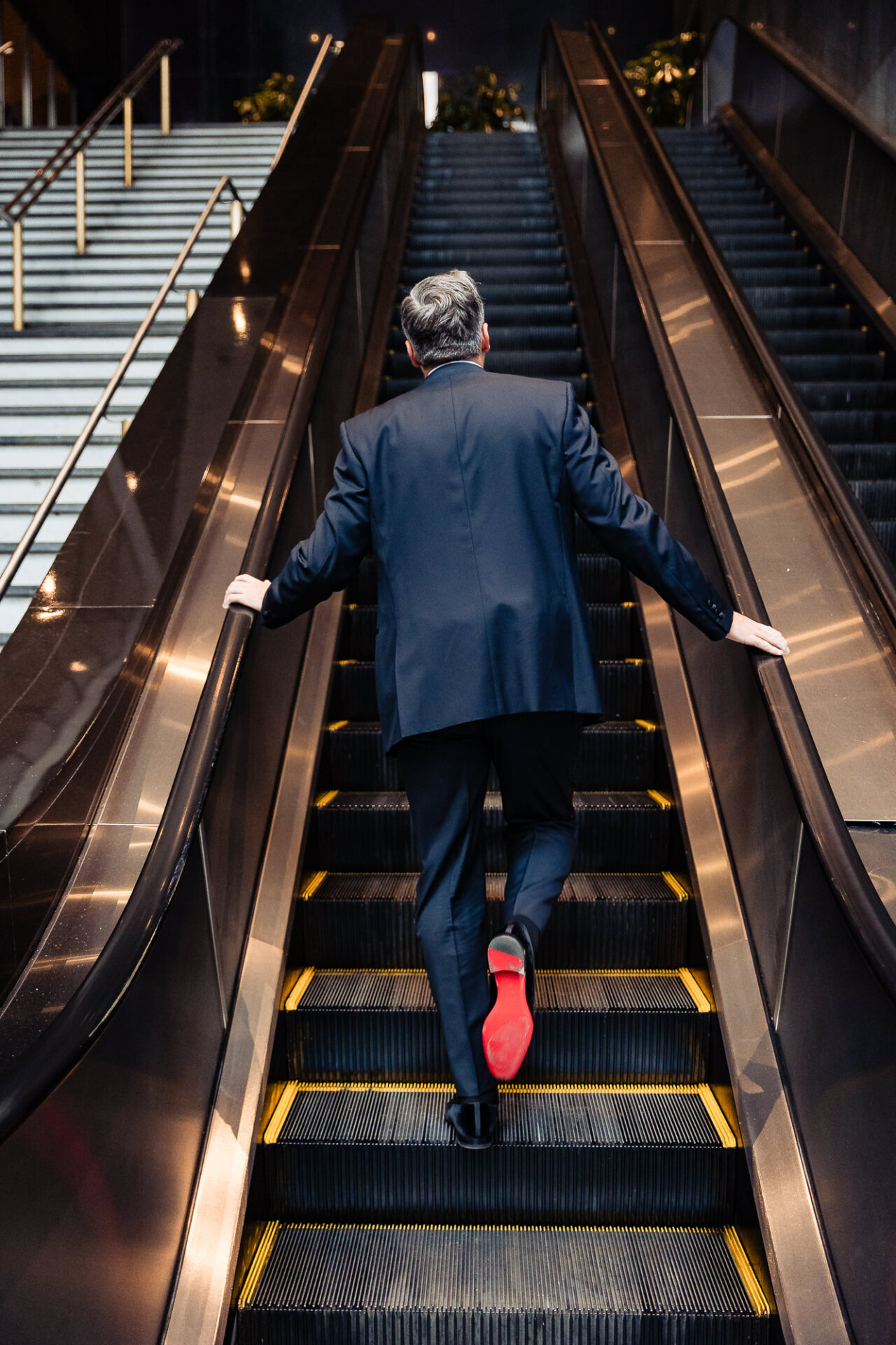 Groom walking up an escalator, holding the handrail with red-soled shoes visible