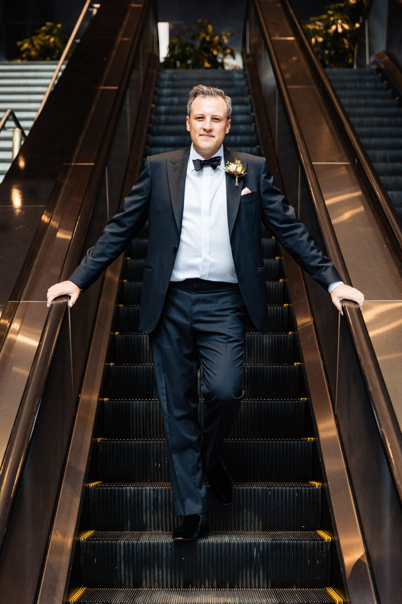 Groom descending an escalator in a tuxedo, hands on the handrails