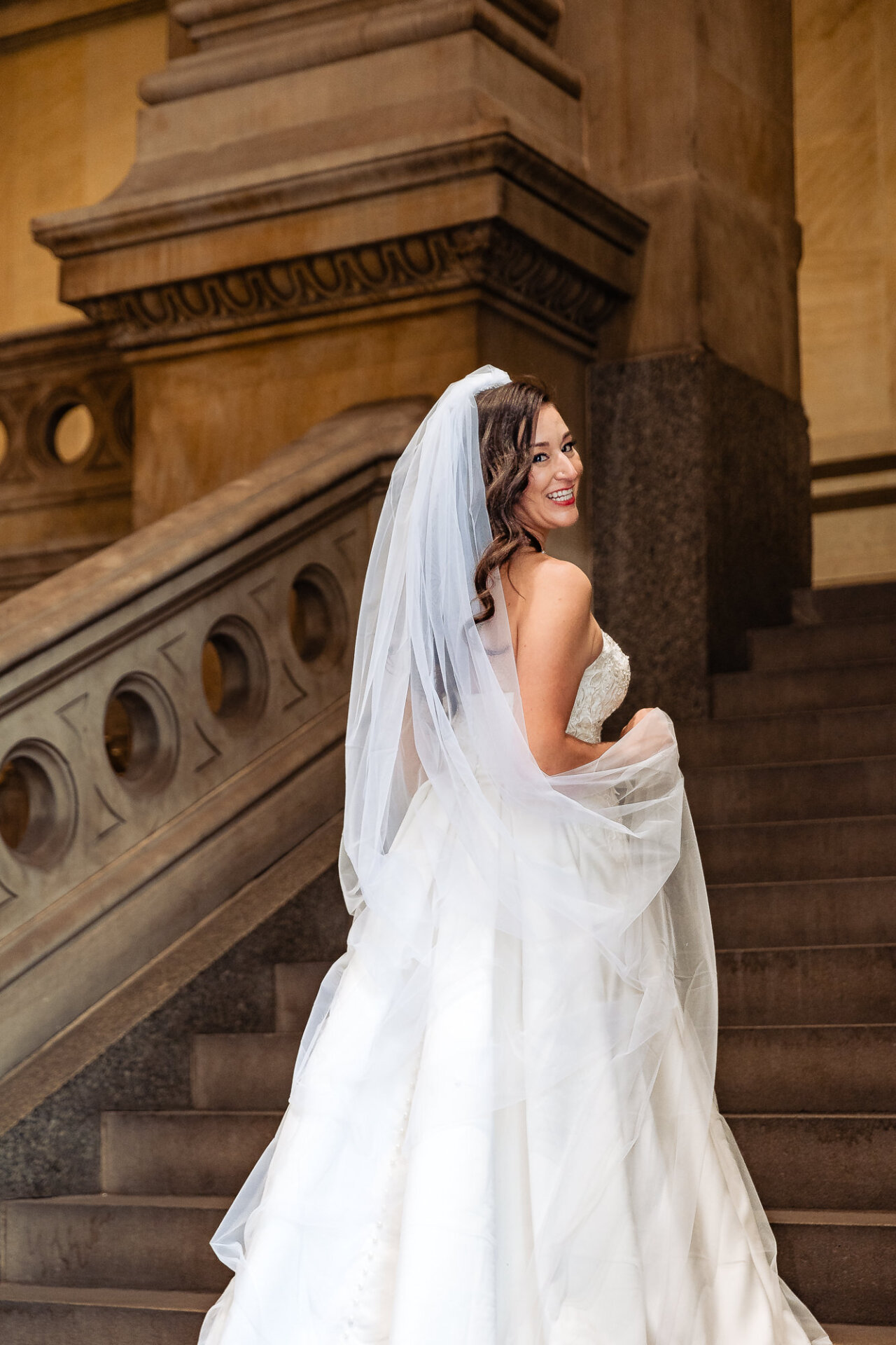 Bride standing on stone steps, smiling with her veil draped over her shoulder