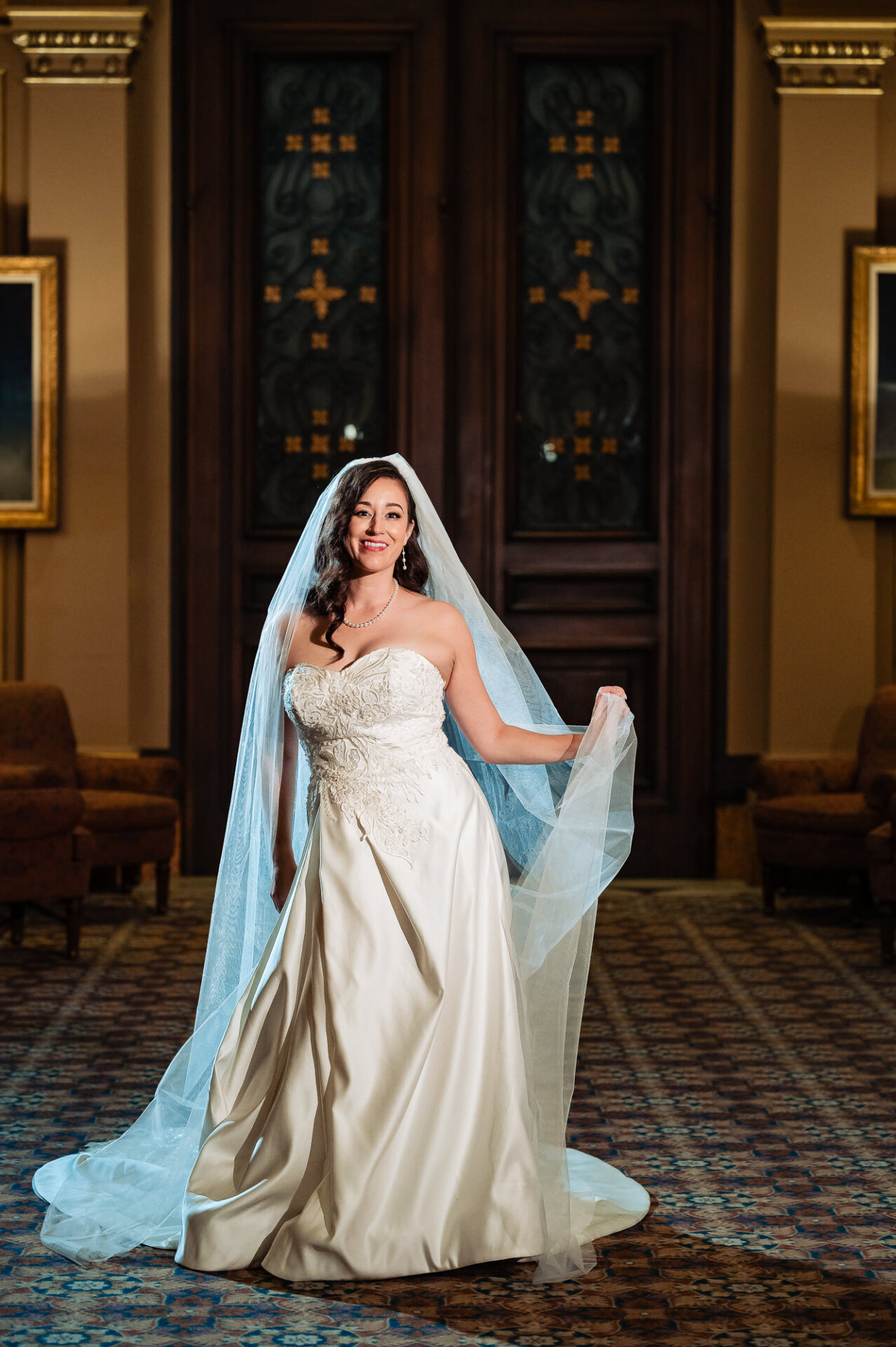 Bride smiling gently, standing in front of large wooden doors with her veil draped over her shoulders