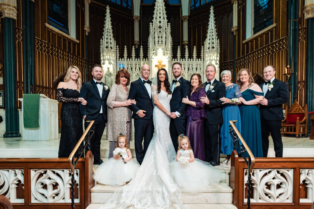 Bride and groom standing with a larger group of family and bridal party members at the altar, with flower girls sitting gracefully at the base of the steps.