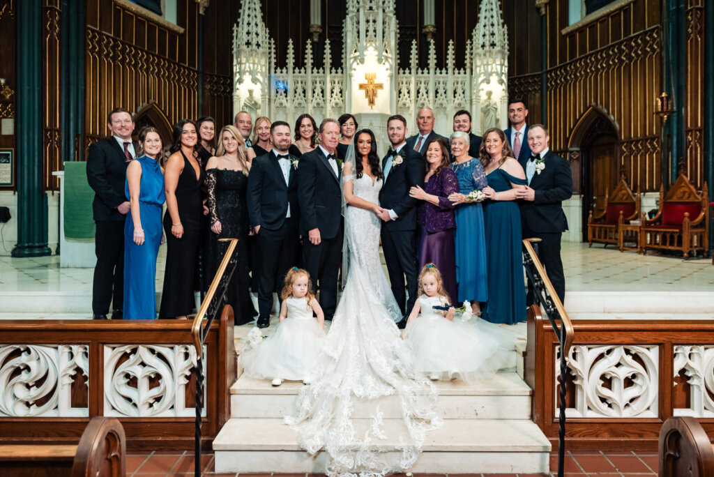 Full bridal party and family members gathered around the bride and groom at the altar, with flower girls sitting in the foreground and everyone smiling warmly