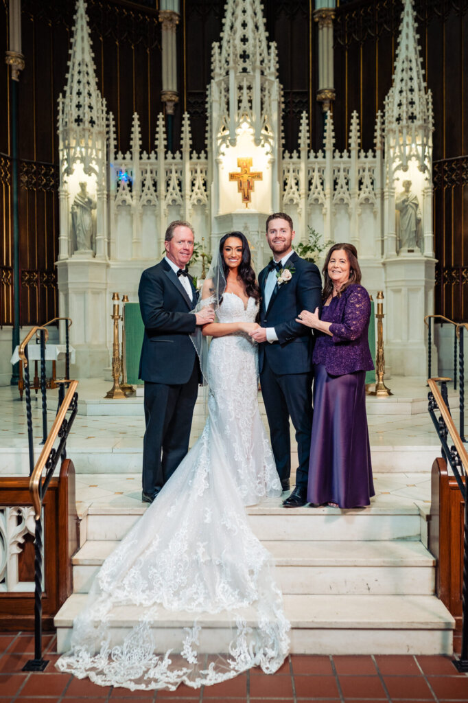 Bride and groom standing at the altar with the groom’s parents, holding hands and smiling, framed by the church’s intricate design.