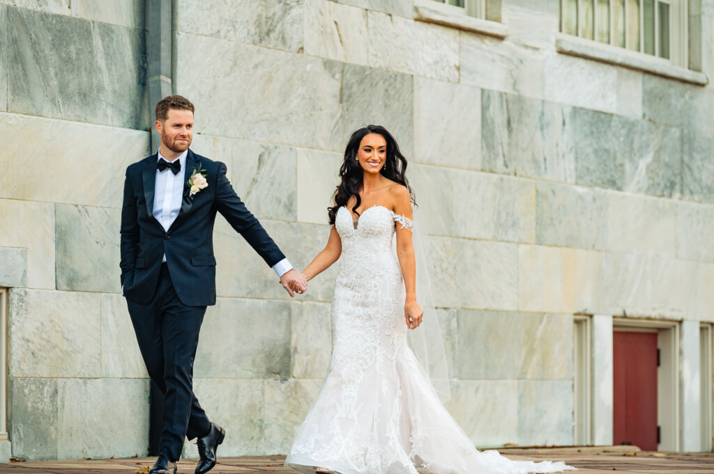 Bride and groom walking hand in hand in front of a stone wall, with her gown and veil flowing gracefully