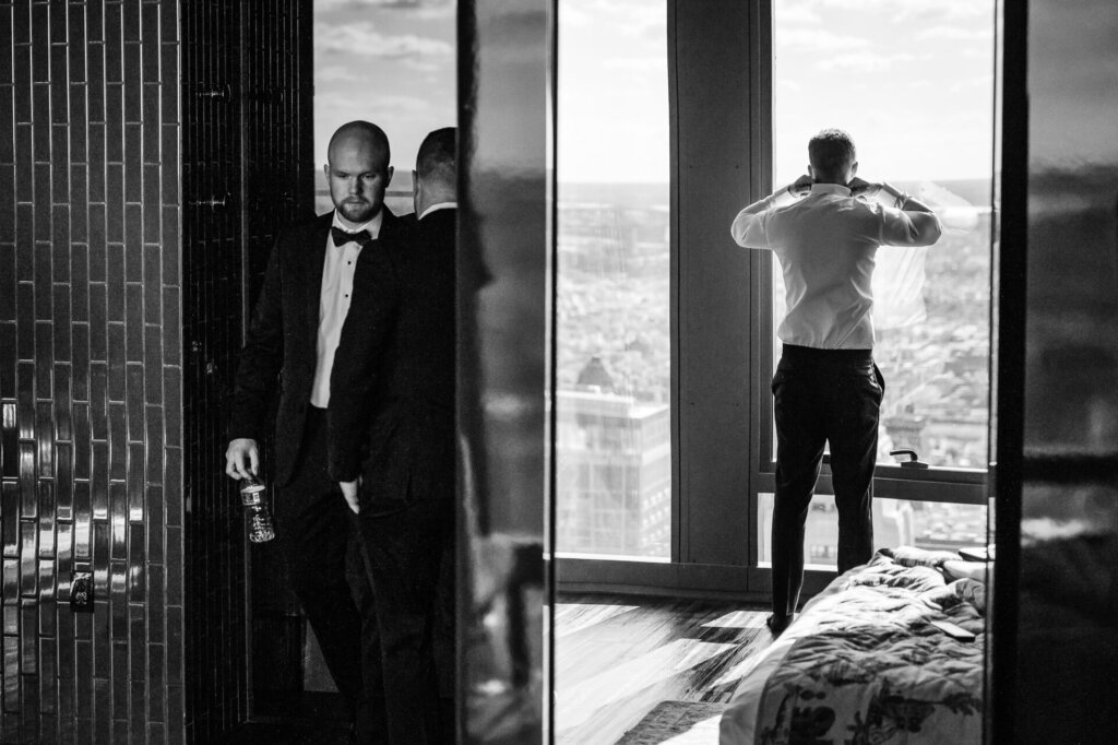 Black and white image of a groom standing by a large window, gazing outside while wearing a tuxedo