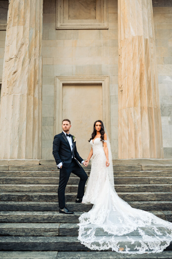 Bride and groom standing together on the wide stone steps of a historic building, holding hands with her lace train cascading beautifully behind