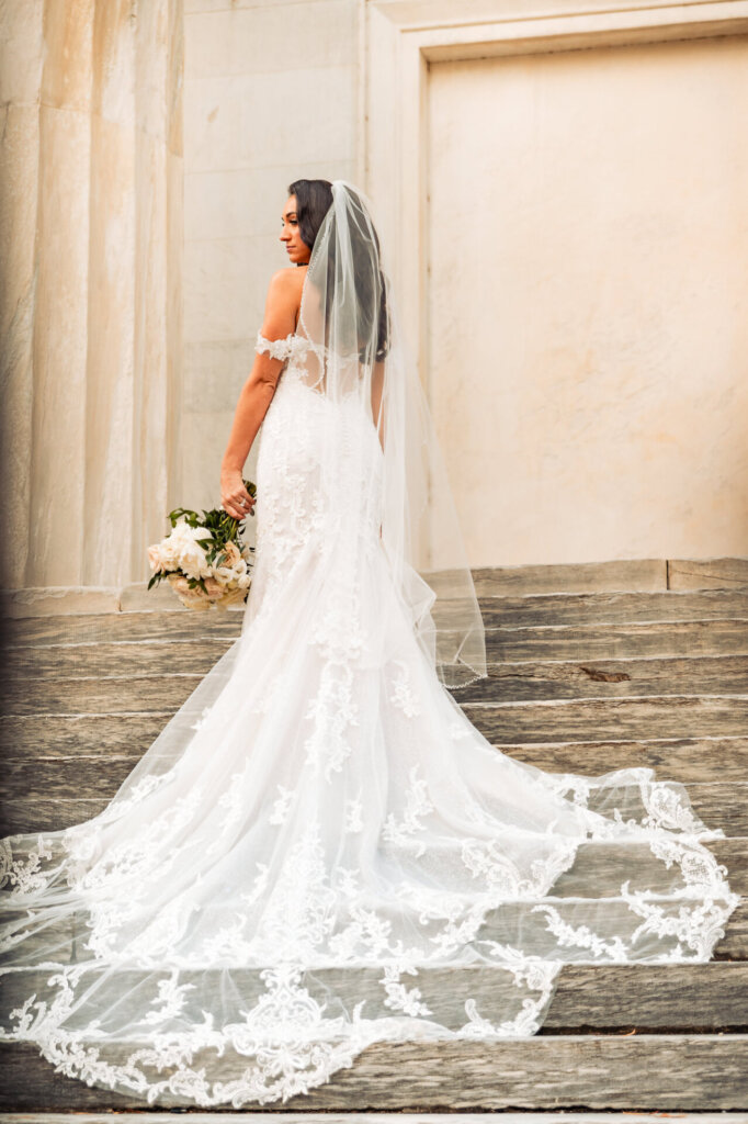 Bride posing gracefully in front of a stone wall, showcasing the train of her lace wedding gown, holding a bouquet in soft sunlight.