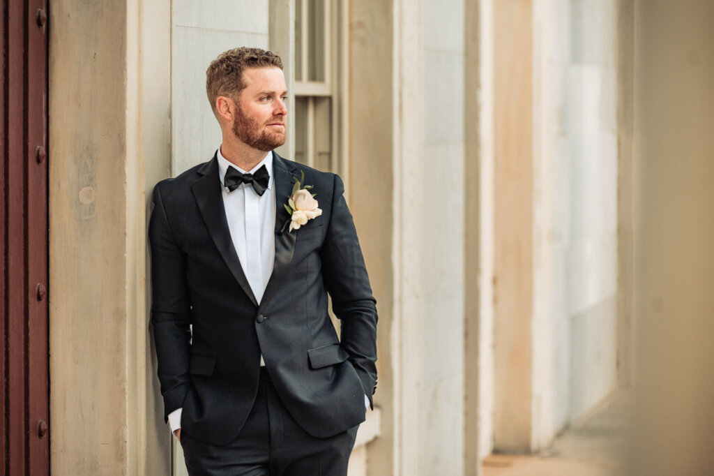 Groom standing in front of a large columned building, dressed in a sharp black tuxedo with a boutonniere, gazing confidently into the distance.