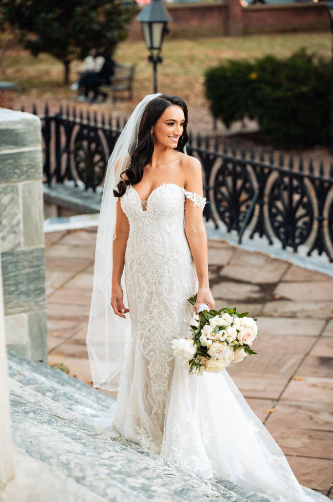 Bride smiling and looking to the side while standing on stone steps near a decorative iron railing, her lace gown glowing in soft natural light.