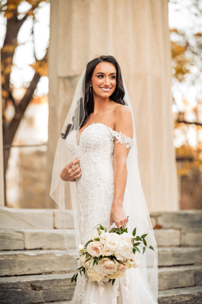 Bride smiling with a soft expression while holding a bouquet, standing in front of grand stone columns, her lace dress highlighting delicate off-the-shoulder details.