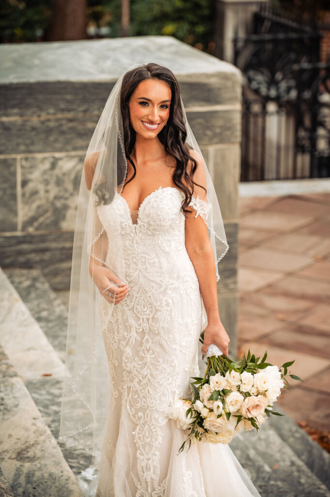 Bride standing on a stone staircase, holding her bouquet, her long lace train displayed elegantly