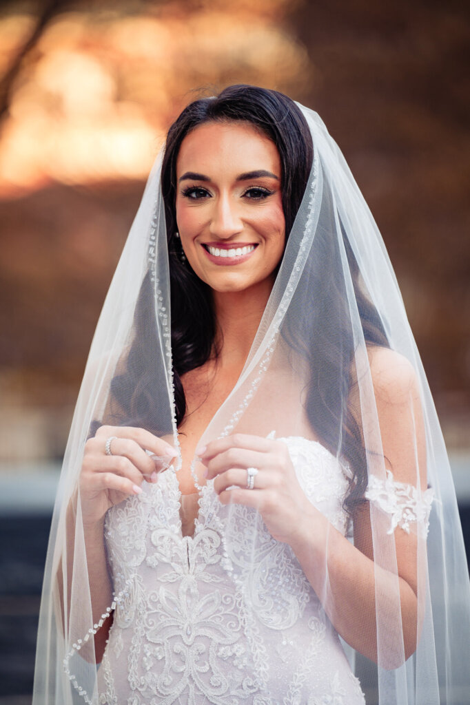 Bride holding her veil gently, smiling softly, with golden light accentuating her lace gown and cascading curls