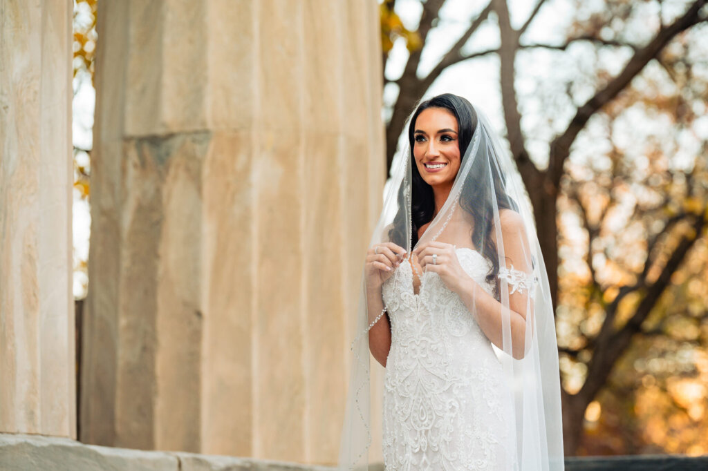 Bride smiling under her veil, holding it delicately with her hands, standing in front of a stone column with autumn trees in the background.