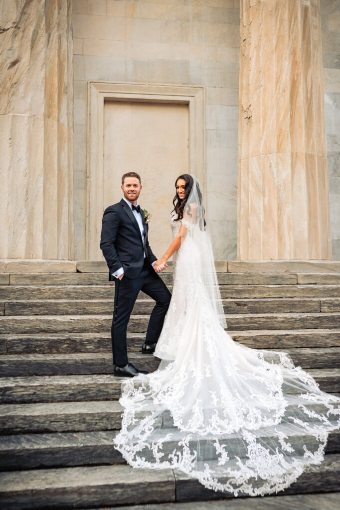 Bride and groom standing on the grand steps of a historic building, holding hands, with the bride’s lace train flowing elegantly down the stairs.
