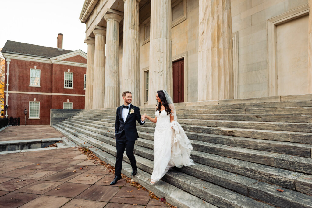 Bride and groom walking hand in hand down the steps of a historic building, with her lace train flowing behind her.