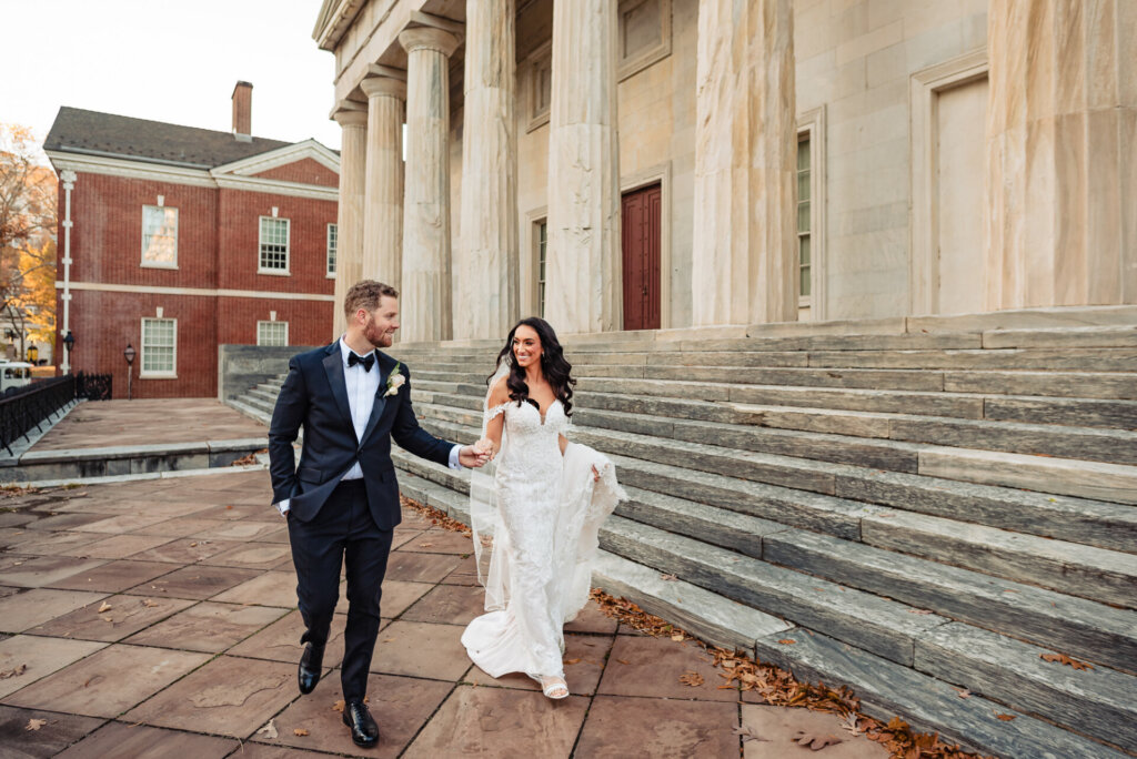 Bride and groom laughing together while descending the steps of a grand stone building, surrounded by historic architecture