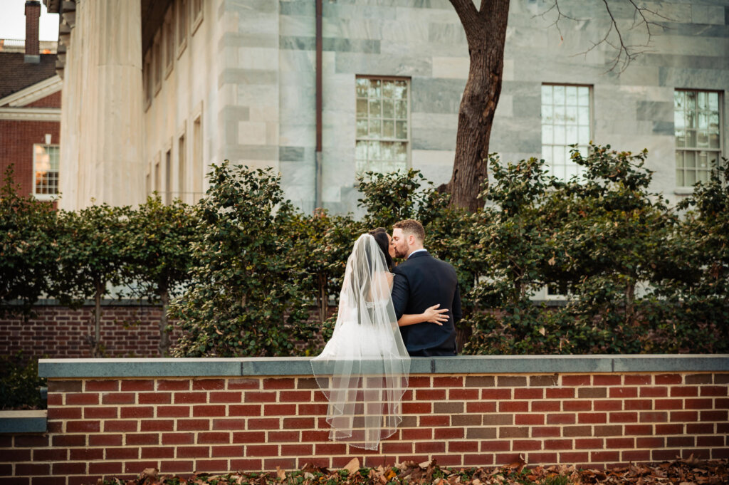 Bride and groom standing close together, sharing a kiss by a brick wall adorned with greenery.
