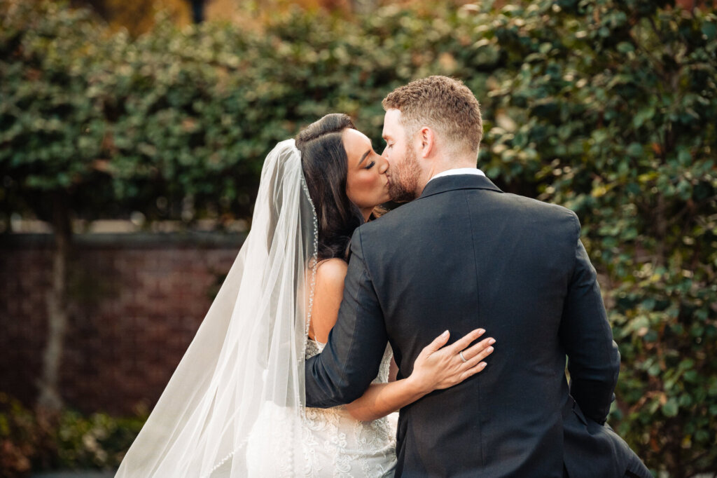 Bride and groom sharing a kiss in front of a brick wall with greenery, their arms wrapped around each other.