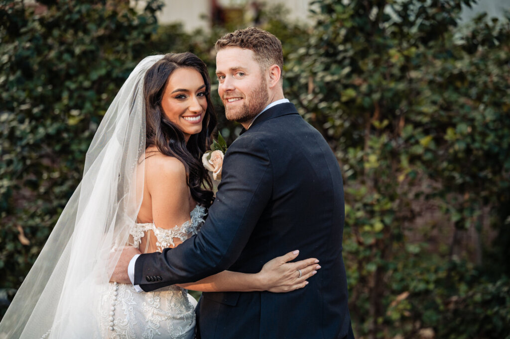Bride and groom side by side, smiling at the camera with lush green hedges behind them.