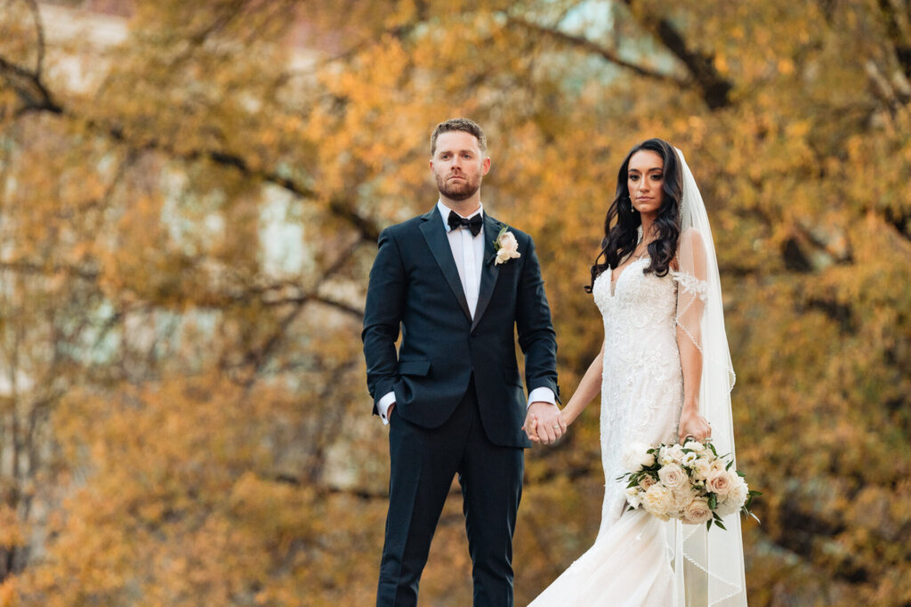 Bride and groom posing together under fall foliage, the groom standing confidently while the bride holds a cascading bouquet.